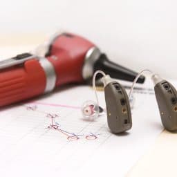 A pair of modern hearing aids on a medical chart showing a hearing loss of high frequencies. An otoscope lies in the background.