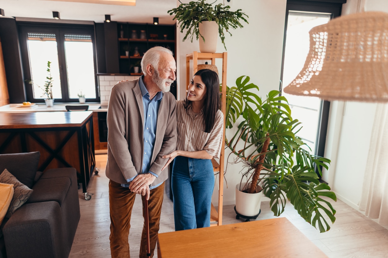 Young woman assisting elderly man with walking stick at home.