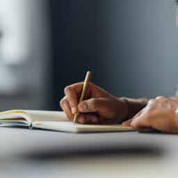 Person wearing a brown sweater writing in their journal on a table.