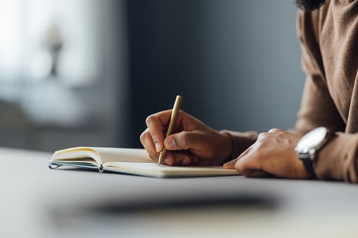 Person wearing a brown sweater writing in their journal on a table.