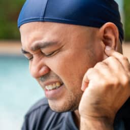 A person wearing a swim cap pinches their ear with a grimace, suggesting ear pain or discomfort after swimming.